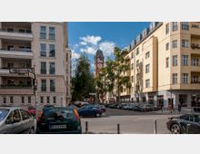 Straßenszene in einer Stadt mit mehrstöckigen Wohnhäusern aus hellem Putz und Balkonen, sowie parkenden Autos am Straßenrand. Im Hintergrund ragt ein rotes Backsteingebäude mit einer Turmuhr und spitzem Dach über Bäume empor.