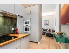 Offener Wohnbereich mit moderner Küche: helle, grifflose Schränke, Holzarbeitsplatte und grüne Fliesenrückwand mit eingebautem Ofen und Mikrowelle. Dahinter schließt ein kleines Wohnzimmer mit grauem Ecksofa an; rechts ein Glas-Wandregal und gerahmte Bilder an weißen Wänden über hellem Holzboden.