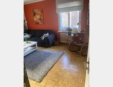 Wohnzimmer mit orangener Wand, einem dunkelblauen Sofa, einem weißen Couchtisch und einem grauen Teppich auf einem Parkettboden. Ein Fenster mit Vorhang lässt Tageslicht herein, daneben stehen ein kleiner Holztisch mit Dekorationen und ein Holz-Schaukelstuhl.