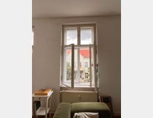 Ein heller Raum mit einem großen Doppelfenster, das auf einen Innenhof mit Wohngebäuden und Bäumen blickt. Vor dem Fenster befinden sich eine Heizung, ein grünes Sofa und ein kleiner Beistelltisch.