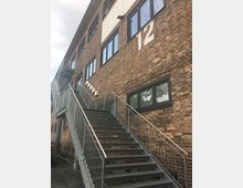 Backstein-Gebäude mit außenliegender Metalltreppe, die zu den oberen Stockwerken führt. Große Fenster und die Zahl "12" sind auf der Fassade sichtbar.