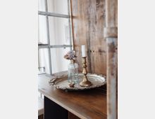 Ein dekoratives Stillleben auf einem Holzregal mit einer silbernen, reich verzierten Metallplatte, auf der ein goldener Kerzenhalter mit einer weißen Kerze, eine kleine Glasflasche mit getrockneten Blumen und ein kleiner Keramikgegenstand stehen. Im Hintergrund sind eine hölzerne Trennwand und ein Metallrahmen zu sehen.