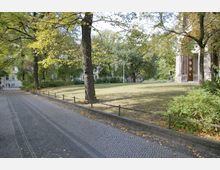 Gepflasterter Weg mit niedriger Metallabsperrung, daneben eine grüne Rasenfläche und mehrere Bäume. Im Hintergrund sind Büsche, Laternen und ein historisches Gebäude mit Säulen und Holzportal sichtbar.