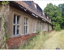 Ein verlassenes, einstöckiges Gebäude mit bröckelndem Putz, zersprungenen Fenstern und einem roten Ziegelsockel. Das Dach ist mit beschädigten Dachziegeln gedeckt, und die Umgebung ist von überwucherndem Gras und Bäumen geprägt.