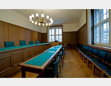 Ein Gerichtssaal mit Holzvertäfelung, einem großen Kronleuchter und Tischen mit grünen Filzauflagen. Die Sitzreihen entlang der Wände bestehen aus Stühlen mit dunkelgrün gepolsterten Sitzflächen, und Tageslicht fällt durch große Sprossenfenster ein.