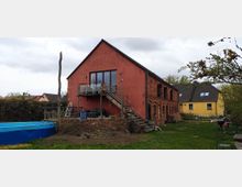Rotes Backsteinhaus mit zwei Etagen, teilweise verputzt, und einer Holztreppe, die zu einem Balkon führt. Der Garten im Vordergrund enthält einen runden, blauen Pool und vereinzelt angepflanzte Sträucher.
