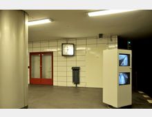 U-Bahn-Station mit weiß gefliesten Wänden, einer roten Doppeltür, einer Wanduhr und einer Überwachungssäule mit zwei Monitoren, die den Bahnsteig zeigen. Im Vordergrund eine silberne Säule und eine Mülltonne an der Wand befestigt.