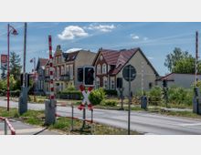 Bahnübergang mit geschlossenen Schranken und Warnsignalen, im Hintergrund Mehrfamilienhäuser mit Satteldächern und teilweise Fachwerkfassade. Die Umgebung ist von Grünflächen und Straßenlaternen mit roten Stangen geprägt.