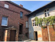 Zweistöckige Backstein- und Putzgebäude, verbunden durch einen Innenhof. Links ein rotes Backsteinhaus mit kleinen Fenstern und einer Treppe, rechts ein verputztes Gebäude mit weißen Fenstern, Holztüren und Weinranken an der Fassade.