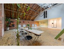 Industrie-inspiriertes Loft mit Backsteinwänden, Holzbalkendecke und Glasfenstern. Die Einrichtung umfasst einen langen Tisch mit Drahtstühlen, Hängepflanzen und eine offene Küche auf der rechten Seite.