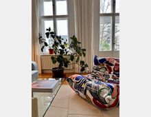 Wohnzimmer mit großen, weißen Sprossenfenstern, langen Vorhängen und einer Zimmerpflanze in einem braunen Topf. Im Vordergrund ein bunt gemusterter Sessel, ein Glas-Couchtisch und ein Teppich auf einem Holzboden.