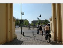 Blick durch ein großes gelbes Tor auf eine Straßenkreuzung mit Fußgängern, einem Radfahrer und mehreren Ampeln. Im Hintergrund sind historische Gebäude und grüne Bäume zu sehen.