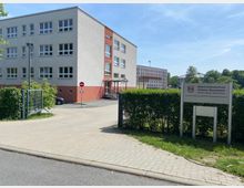 Gebäudekomplex des Oberstufenzentrums Dahme-Spreewald am Standort Königs Wusterhausen, bestehend aus einem modernen, mehrstöckigen, grau-roten Gebäude mit zahlreichen Fenstern; im Vordergrund ein gepflasterter Gehweg, ein Parkplatz sowie ein Schild mit der Aufschrift der Schule.