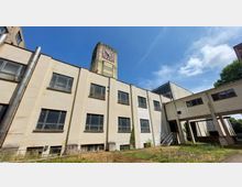 Außenansicht eines alten Industriegebäudes mit verwitterter Fassade, rechteckigen Fenstern und einem angrenzenden Uhrturm. Im Vordergrund befindet sich eine verwilderte Grünfläche, während der Himmel klar und blau ist.