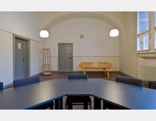 Schlichter Konferenzraum mit einer grau-schwarzen Tischecke und gepolsterten Stühlen im Vordergrund, grauen Türen an der Rückwand, einer Holzbank, einem freistehenden Holz-Kleiderständer und einer Wanduhr. Große Fenster auf der rechten Seite lassen Tageslicht herein.