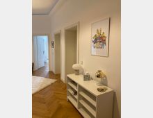 Heller Flur in einer Wohnung mit weiß gestrichenen Wänden, einer Holzdielenboden im Fischgrätmuster und mehreren Türen. Rechts steht ein weißes Regal mit Dekoration, darunter eine Tischlampe, eine Dose und ein Glasgefäß, an der Wand darüber hängt ein buntes Bild.