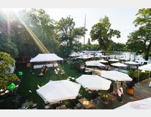 Ein Außenbereich eines Restaurants oder Biergartens an einem Fluss mit weißen Sonnenschirmen, Sitzplätzen aus Holz und einem weißen Zelt für ein Buffet; im Hintergrund sind Bäume und das Wasser sichtbar, mit Stegen und Booten am Ufer.