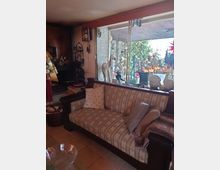 Gemütlicher Wohnraum mit einem gestreiften Sofa, dekoriert mit Kissen und einer Decke, vor einer großen Glasfensterfront. Im Hintergrund ist ein Außenbereich mit Pflanzen und Steinmauern sichtbar; nahe der Fensterbank stehen kleine Dekorationsgegenstände wie Holzspielzeug.