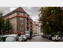 Straßenszene mit gepflasterter Fahrbahn, gesäumt von parkenden Autos. Links steht ein mehrstöckiges Wohngebäude mit brauner Fassade und roten Dachziegeln, während rechts Bäume am Straßenrand zu sehen sind.