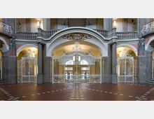 Historischer Innenraum mit verziertem, weißem Metallgittertor vor einem Eingangsbereich. Umgeben von Rundbögen, Säulen und kunstvoll bemalten Wänden mit geschwungenen Stilmustern; der Boden besteht aus roten Fliesen mit einem Muster aus weißen Quadraten.