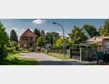 Wohnstraße in einer Vorstadt mit Einfamilienhäusern und Gärten, darunter ein hellbraunes Haus mit rotem Dach. Die Straße ist schmal, von Bäumen und Zäunen gesäumt, und ein einzelner Laternenmast steht am Straßenrand.