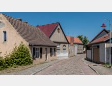 Eine gepflasterte Straße in einem Dorf mit kleinen Wohnhäusern, die teils renoviert und teils verfallen wirken. Die Häuser haben rote und braune Ziegeldächer, vereinzelt Fensterläden, und rechts ist eine Straßenlaterne mit rotem Lampenschirm zu sehen.