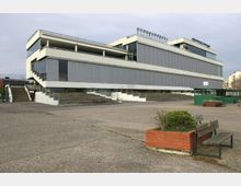 Modernes, rechteckiges Gebäude mit mehreren Etagen und grauen Lamellenfassaden, flankiert von symmetrischen Außentreppen. Im Vordergrund befinden sich eine gepflasterte Fläche, Bänke und ein kleiner gemauerter Blumenkübel.