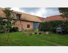 Ein grasbewachsener Garten mit einer Sitzgruppe aus Baumstämmen um eine Feuerstelle. Im Hintergrund steht ein Backsteinhaus mit Holztür und Satteldach, umgeben von Büschen und einem Grill auf der rechten Seite.