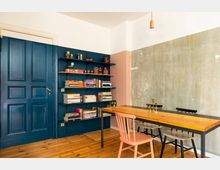 Esszimmer mit Holzboden, einem langen Tisch mit schwarzem Metallrahmen und gemischten Stühlen, darunter ein rosa Stuhl. Links ein dunkelblaues Regal voller Bücher und Dekorationsgegenstände vor einer blau gestrichenen Tür; die rechte Wand ist teilweise verputzt und zeigt dezente Patina.