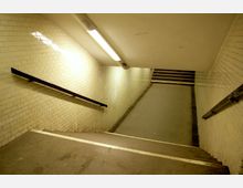 Treppe in einem U-Bahn- oder Unterführungstunnel mit beigen Fliesenwänden und einfacher Deckenbeleuchtung. An den Wänden befinden sich Handläufe, und am Ende der Treppe schließen weitere Stufen an.