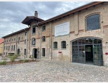 Backsteinbau mit historischem Charakter, großer Glaseingang und mehreren kleinen Fenstern, davor ein mit Pflastersteinen befestigter Hof mit Sitzgelegenheiten und Pflanzen. Ein Schild mit der Aufschrift „Storchenhof Blumenow“ ist an der Fassade angebracht.
