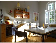 Helle, gemütliche Küche mit Holzfußboden, einer Küchenzeile aus Holz, offenen Regalen mit Geschirr und zwei hängenden Lampen. Ein Esstisch mit Stühlen steht vor einem großen Fenster, durch das Sonnenlicht einfällt.