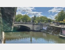 Flussbrücke in einer städtischen Umgebung mit Figurenstatuen auf dem Geländer, flankiert von mit Graffiti bedeckten Betonwänden. Im Hintergrund sind Bäume und ein Kirchturm sowie Fußgänger und Verkehrsampeln zu sehen.