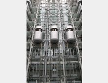 Ein modernes Atrium mit einer hohen Glasfassade, in der drei silberne, runde Aufzüge in transparenten Schächten vertikal verlaufen. Umgeben von Stahl- und Glasplattformen sowie mehreren Ebenen mit Geländern.