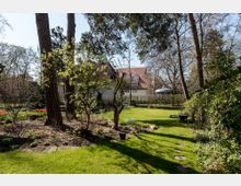 Ein gepflegter Garten mit Rasenfläche, Blumenbeeten und hohen Bäumen; im Hintergrund ein großes Wohnhaus mit rotem Satteldach und Gaubenfenstern. Rechts ist eine kleine, runde Pergola erkennbar, und links blühen rote Tulpen.