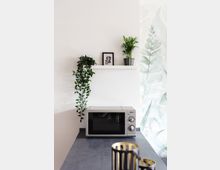 Minimalistische Küchenecke mit einer Mikrowelle auf einer grauen Arbeitsfläche, darüber ein weißes Regal mit zwei kleinen Pflanzen und einem gerahmten Blattmotiv; die Wand ist mit einem dezenten Pflanzenmuster dekoriert.