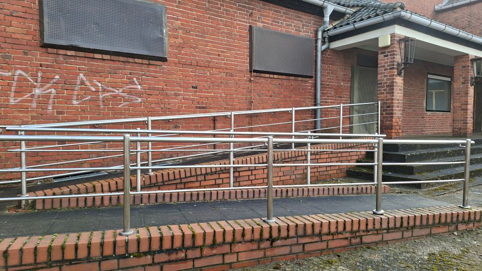 Außenansicht eines Backsteingebäudes mit einer barrierefreien Rampe mit Edelstahlgeländern, die zu einem überdachten Eingang führt. Daneben sind eine kurze Treppe, mehrere abgedeckte Fensteröffnungen und Regenrinnen an der Fassade zu sehen.
