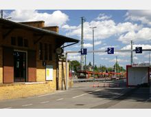 Bahnhofsbereich mit einem historischen Backsteingebäude links, ausgestattet mit braunen Fensterläden und einer analogen Uhr über dem Eingang. Im Hintergrund sind Gleise, Bahnsteige mit blauen Nummernschildern und Strommasten unter einem blauen Himmel mit weißen Wolken zu sehen.
