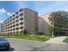 Mehrstöckiges Wohngebäude mit Balkonen auf der linken Seite, in einem moderneren Stil mit pastellfarbenen Wänden. Rechts davon eine Grünfläche mit Bäumen und einem gepflasterten Gehweg, im Vordergrund eine Straße mit parkenden Autos.