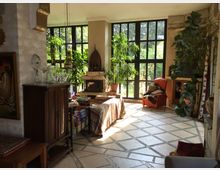 Wohnraum mit großen Fenstern, durch die viel Tageslicht einfällt. Der Raum ist mit Pflanzen, einem Kamin, einem Sessel und Holzmöbeln eingerichtet; der Boden hat ein Muster aus diagonalen Fliesen.