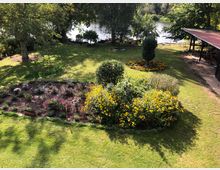 Garten mit gepflegtem Rasen, Blumenbeeten mit bunten Blüten und einzelnen Sträuchern, im Hintergrund ein See mit Bäumen entlang des Ufers. Rechts befindet sich eine überdachte Terrasse mit Sitzmöglichkeiten.