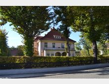 Zweistöckiges Wohnhaus mit hellrosa Fassade und rotem Satteldach, umgeben von hohen Bäumen, einem gepflegten Garten und einer schwarzen Holzzaunbegrenzung. Im Vordergrund befindet sich eine gepflasterte Straße.