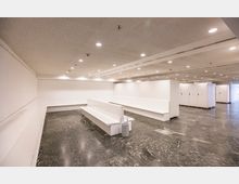 Ein heller, moderner Wartebereich mit weißen Bänken auf einem glänzenden, dunklen Marmorboden. Die Decke ist mit eingebauten Leuchten versehen, und am Rand des Raumes befinden sich weiße Schließfächer.