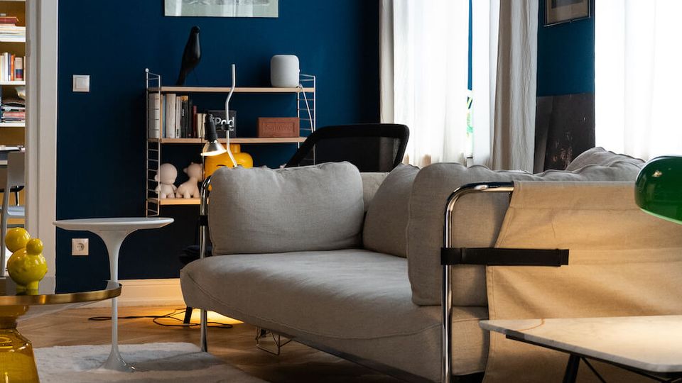 Modern living room with deep blue walls, tall windows covered by sheer white curtains, and a gray sectional sofa on a white rug over herringbone wood flooring. A small bookshelf, side tables, and minimalist framed artwork and lamps add to the contemporary interior.