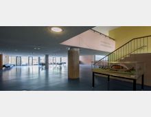 Geräumiges Foyer mit modernem Design, blauen Böden und großen Fenstern, durch die Tageslicht einfällt. Eine Treppe mit Geländer führt nach oben, daneben steht ein architektonisches Modell in einer Glasvitrine, und im Hintergrund befinden sich Sitzgruppen.