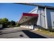 Industriegebäude mit Metallfassade, überdachtem Ladebereich und rotem Dachvorsprung. Im Vordergrund eine asphaltierte Fläche, im Hintergrund Bäume und Vegetation.