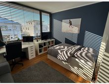 Helles Schlafzimmer mit großen Fenstern und Jalousien, durch die Sonnenlicht und Schattenmuster auf Wände und Möbel fallen. Im Raum befinden sich ein Bett, ein Schreibtisch mit Computer und Bürostuhl, ein kleines Regal mit Fernseher sowie ein grauer Teppich auf dem Holzboden.