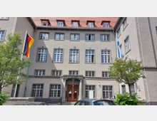 Fassade eines mehrstöckigen, historischen Gebäudes mit symmetrisch angeordneten Fenstern und einem roten Ziegeldach. Vor dem Eingang mit einem steinernen Portal hängen die deutsche und eine blau-weiße Flagge, umgeben von Bäumen und parkenden Autos.