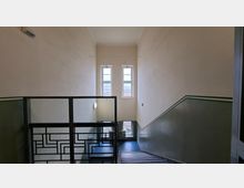 Treppenhaus in einem Mehrfamilienhaus mit hohen, hellen Wänden und einem dunkelgrünen Wandstreifen entlang der Treppe. Ein Metallgeländer mit geometrischem Muster führt hinab zu zwei hohen Fenstern am Treppenabsatz, durch die Tageslicht einfällt.
