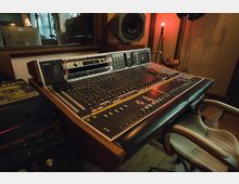 Mischpult in einem Tonstudio mit zahlreichen Reglern, Knöpfen und Schaltern; im Hintergrund sind ein Lautsprecher, eine rote Lampe und eine Glasscheibe zu einem weiteren Raum sichtbar. Ein professioneller Arbeitsstuhl steht vor dem Pult.